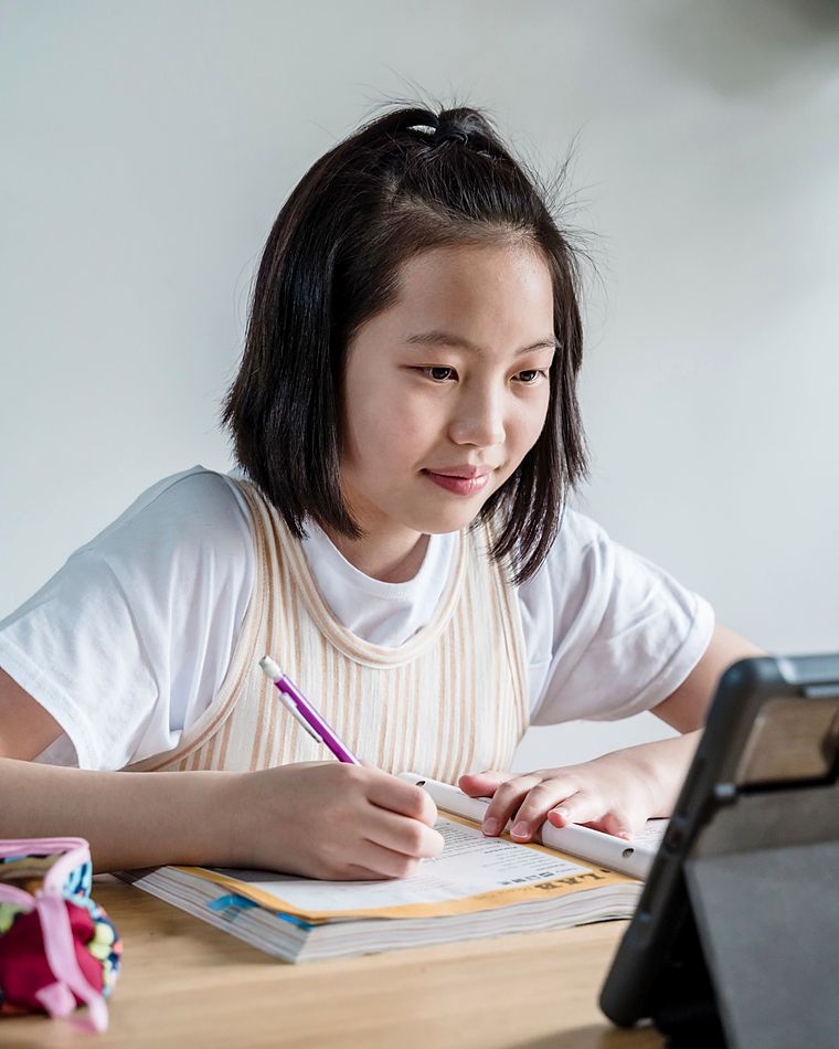 A girl smiles as she writes in her workbook, her tablet open beside her for the lesson